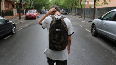 boy wearing white shirt and black shorts carrying backpack standing on black concrete road between vehicles and trees during daytime
