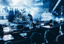 man in black t-shirt and black jeans standing on stage near drum set
